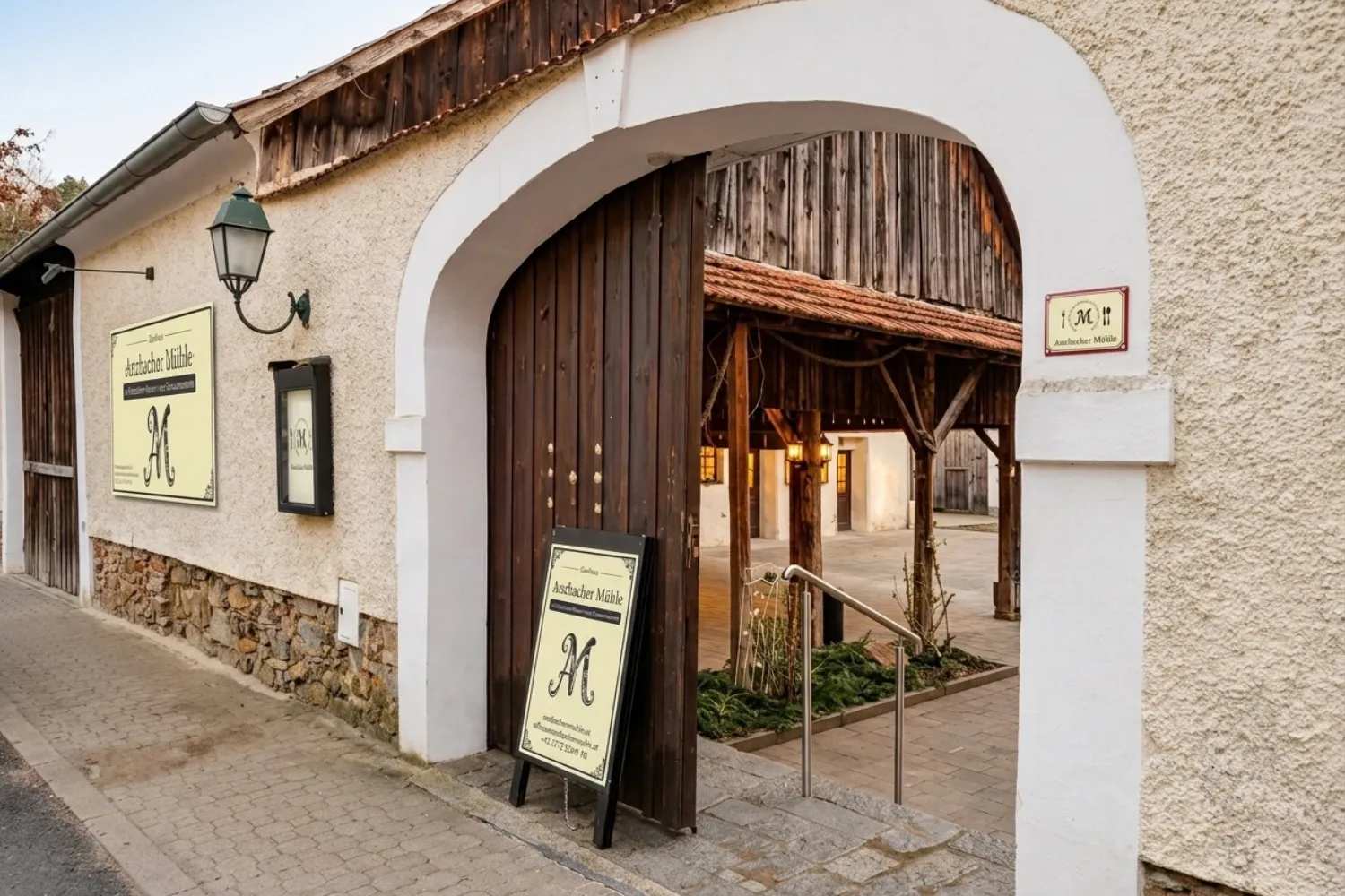 Der Eingang der Anzbacher Mühle: ein weißer Torbogen mit Holztor, Schildern mit dem Mühlen-Wappen und Blick in den Innenhof