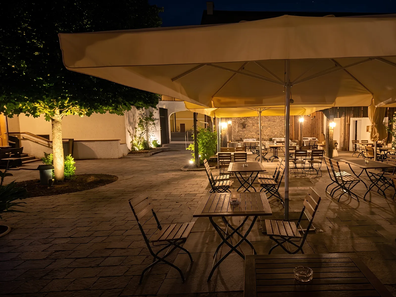 Der Gastgarten im Innenhof der Anzbacher Mühle am Abend, mit Sonnenschirm, Holztischen und beleuchtetem Baum